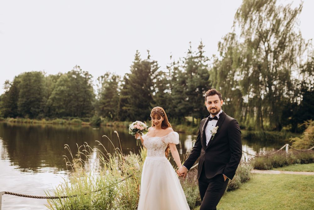 A wedding couple poses together on a manicured lawn beside a tranquil pond surrounded by pine trees.