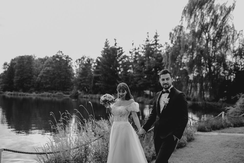 A black and white portrait shows a couple in wedding attire standing by a peaceful lake with evergreen trees.