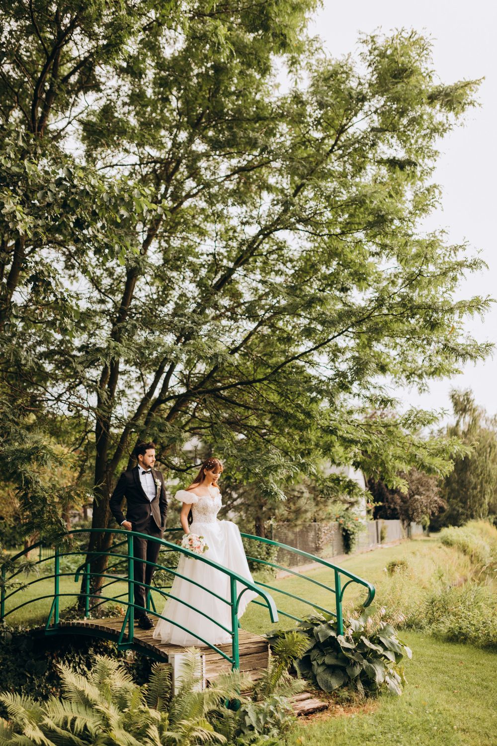 A wedding couple stands on a green bridge in a garden setting with large trees providing natural shade.