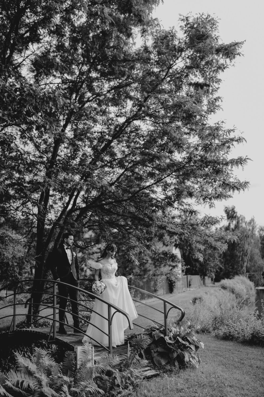 A black and white wedding photo shows a couple standing on a garden bridge surrounded by lush foliage.