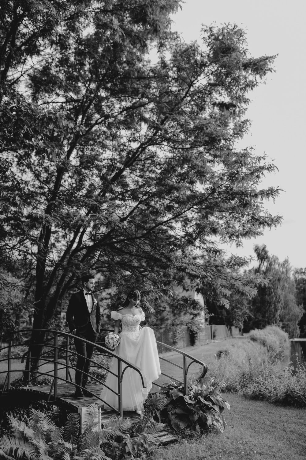 A couple stands on a rustic garden bridge surrounded by lush trees and foliage in a black and white photo.