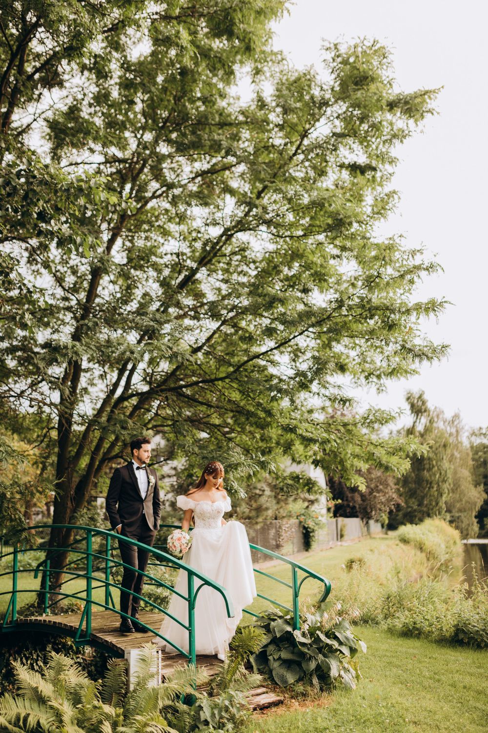 A couple in wedding attire poses on an ornate green bridge in a landscaped garden with towering trees.