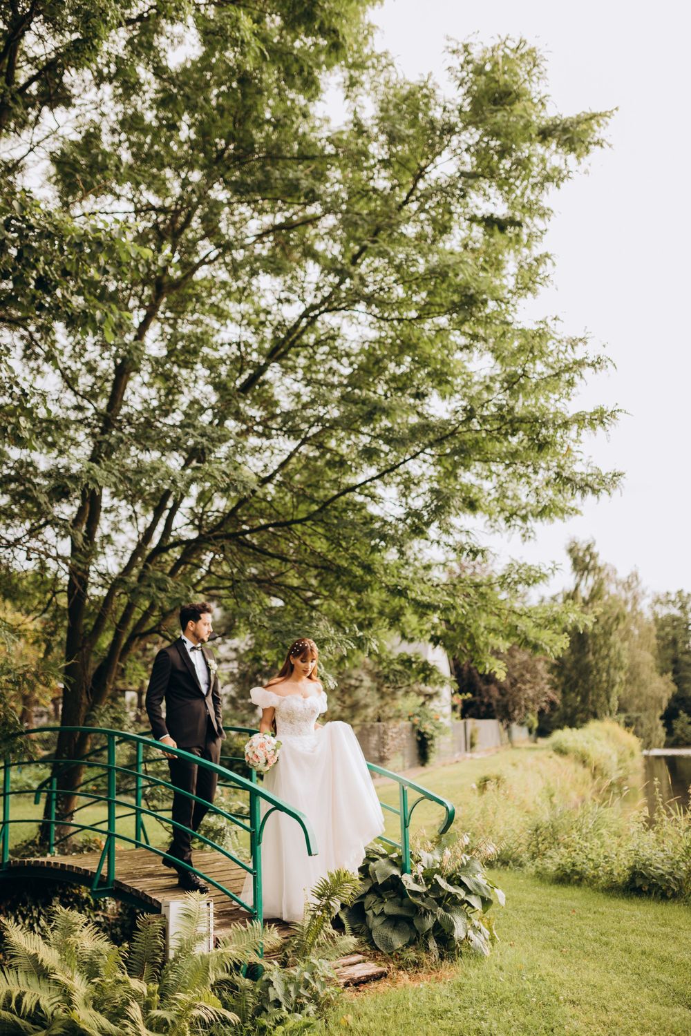 A wedding couple poses on a green metal bridge surrounded by trees and landscaping.