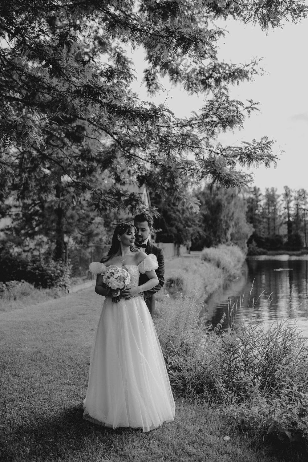A romantic black and white photo of a couple by a serene pond surrounded by trees.