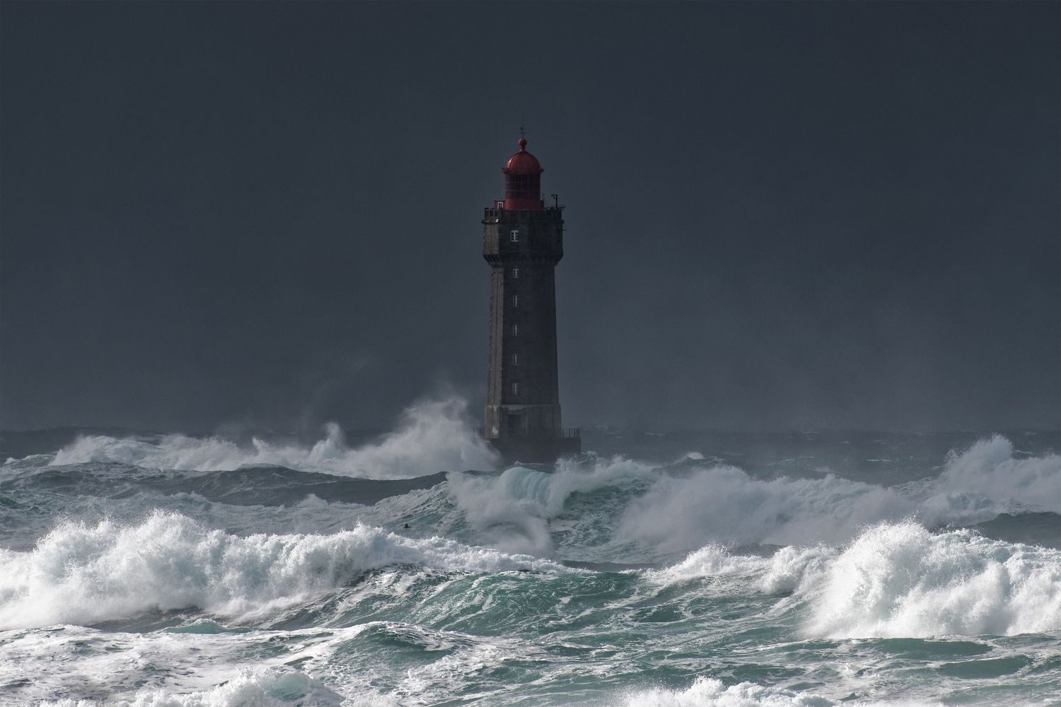 Lighthouse standing against dark stormy seas with large waves crashing around its base.