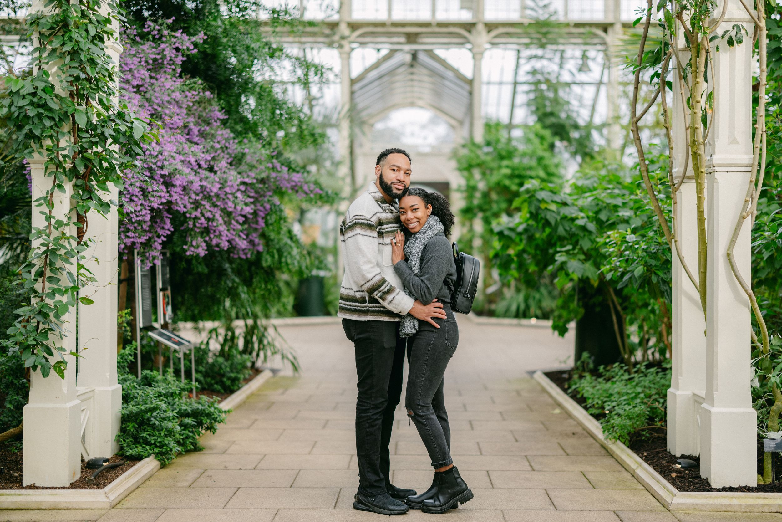 A couple embracing in a greenhouse walkway surrounded by purple flowers and greenery.