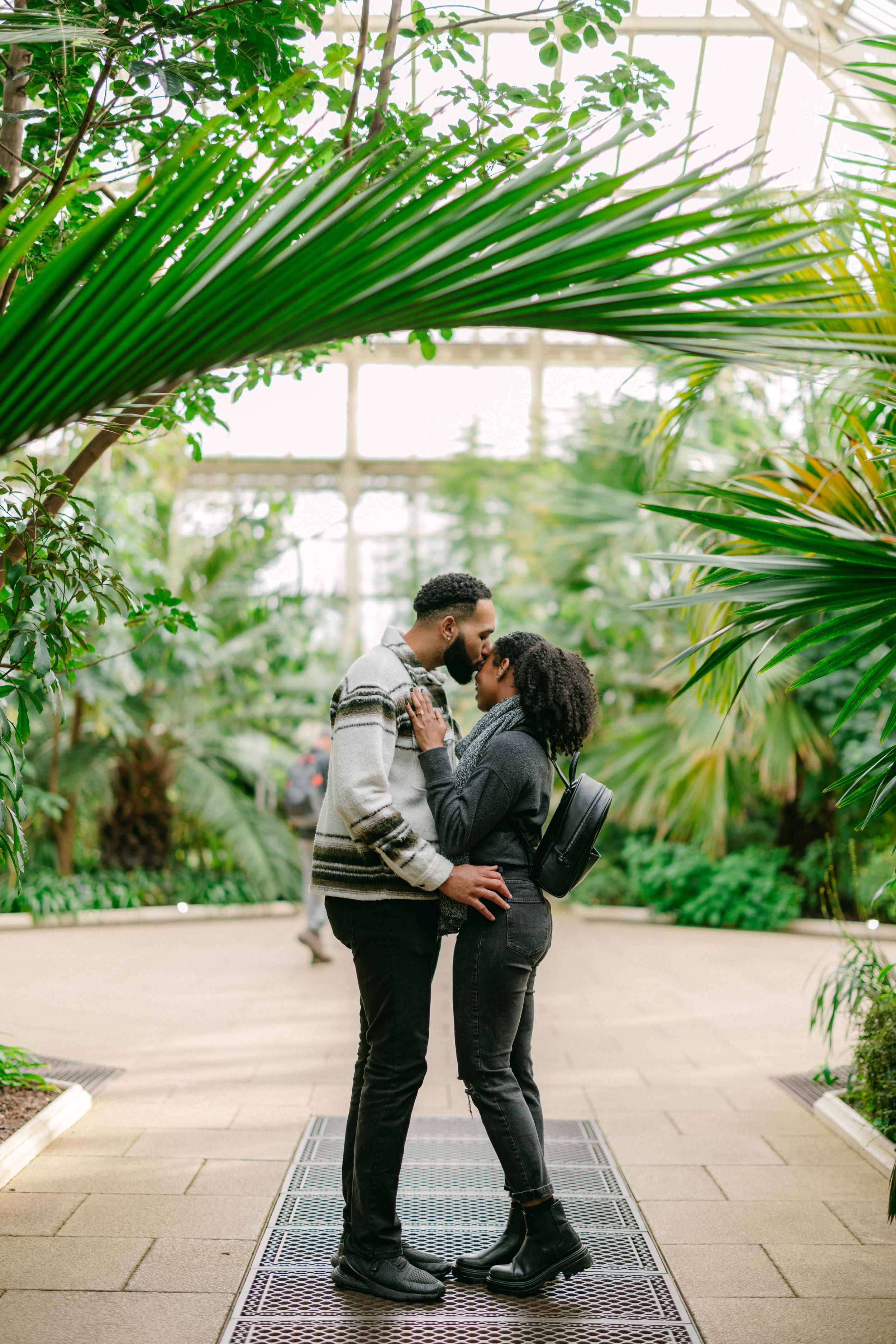 A couple sharing a romantic moment under palm fronds in a greenhouse setting.