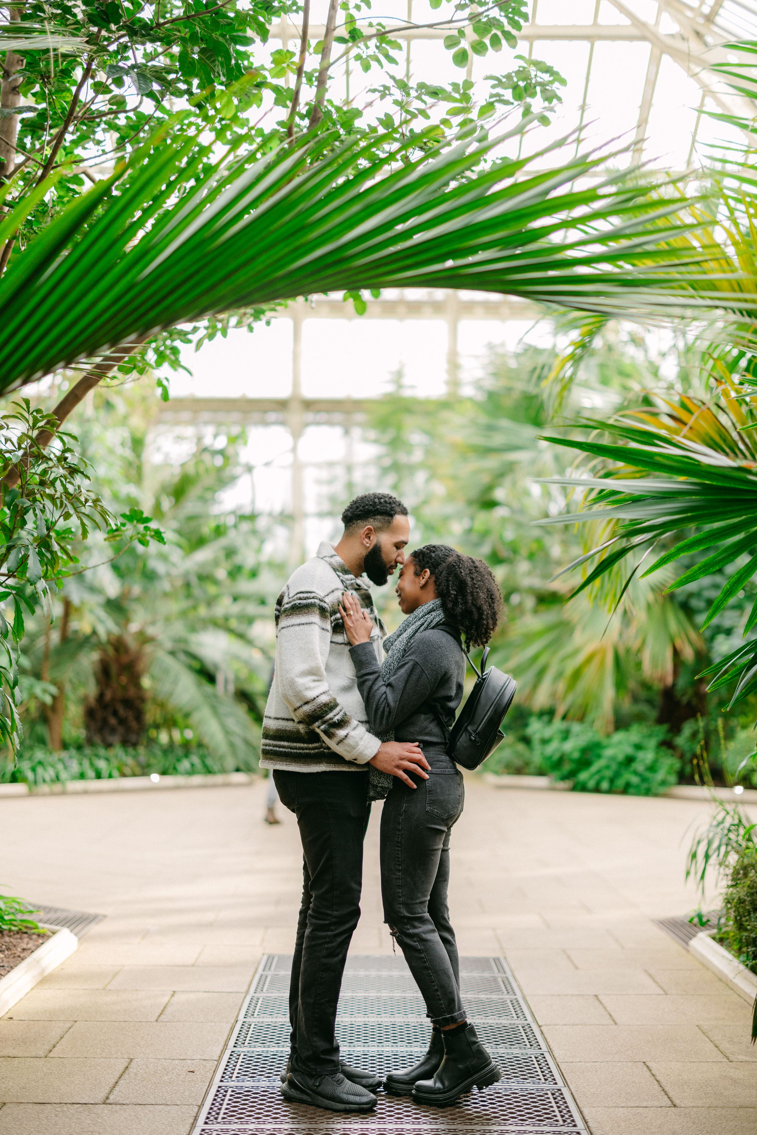 A romantic couple embraces in a lush greenhouse surrounded by tropical palm fronds and natural light.