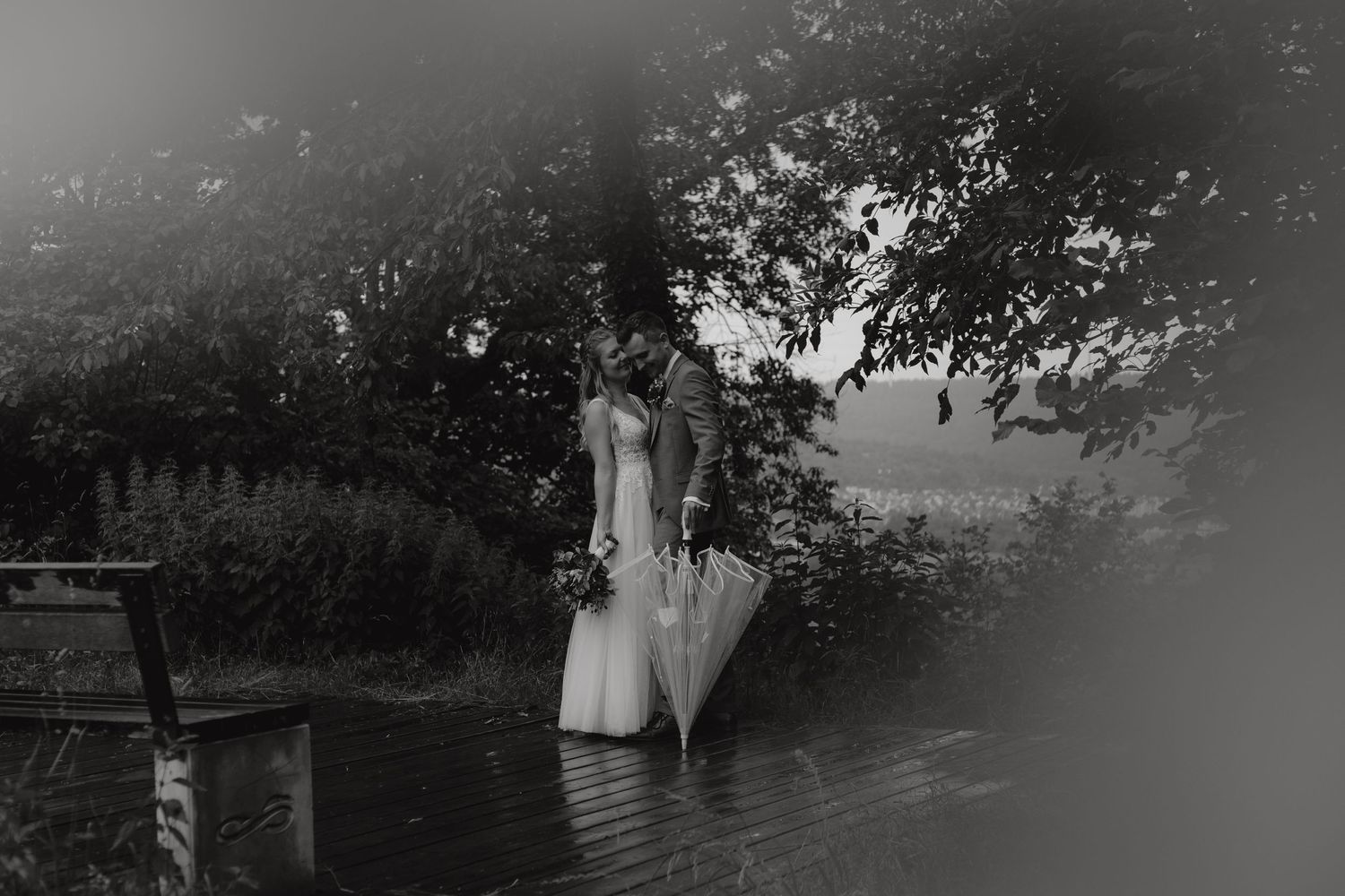 A dreamy black and white photo of a bride standing near water under overhanging trees.