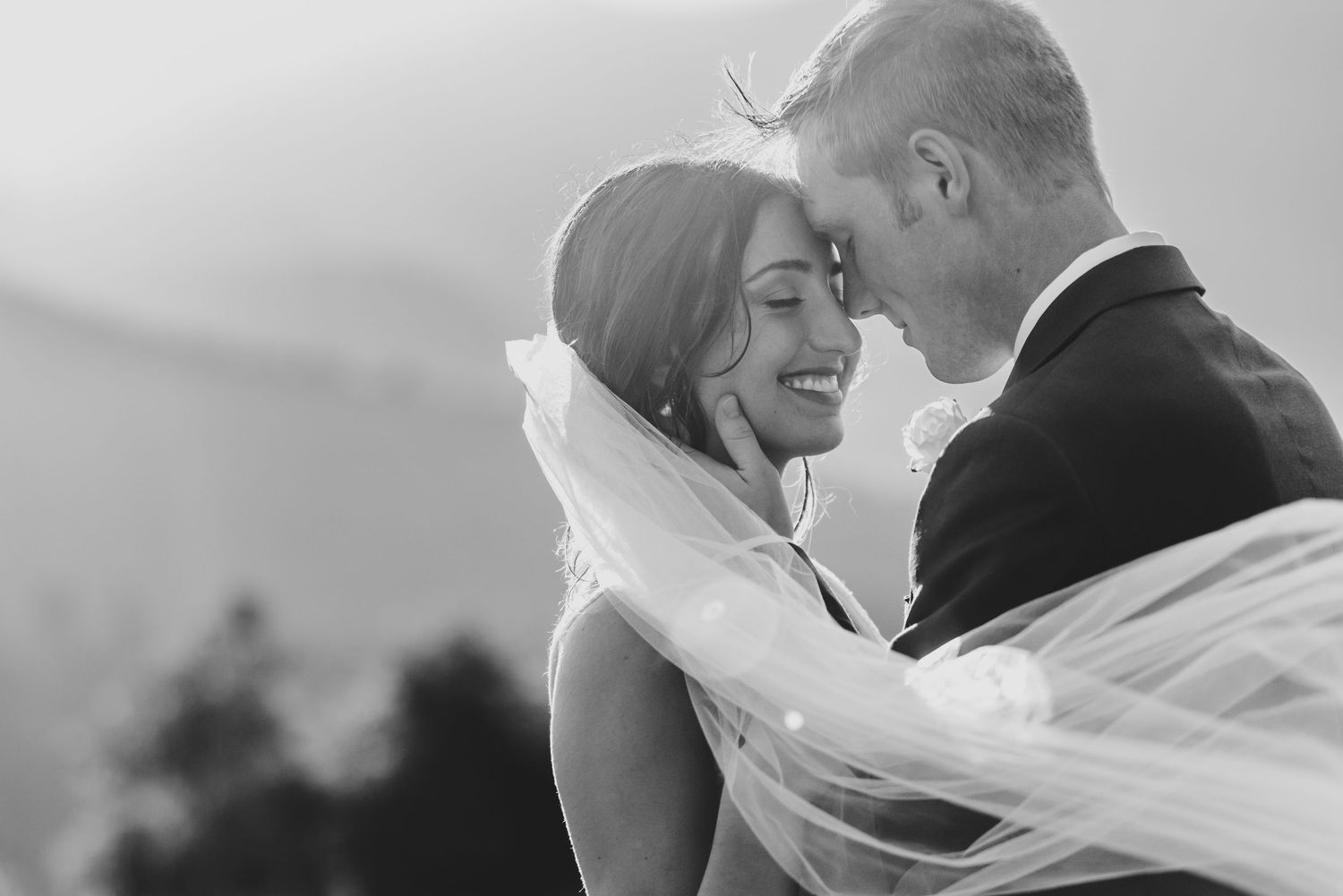 Intimate black and white portrait of couple with flowing veil against mountain backdrop.