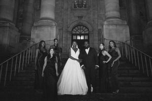 Wedding party in formal attire poses on stone steps of historic building in black and white.