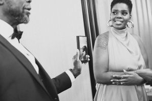 A candid black and white photo showing someone taking a phone picture of a smiling woman in formal attire.