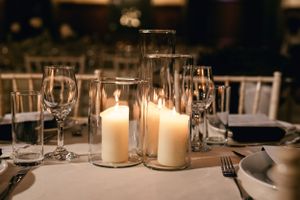 Elegant wedding reception table setting with glowing candles in glass cylinders and wine glasses.