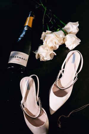 Wedding details featuring white roses, champagne bottle, and elegant bridal shoes on black background.