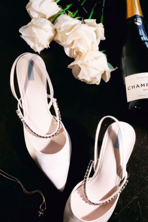 Elegant white bridal shoes with crystal straps displayed with roses and champagne.