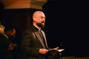 A man in dark suit and glasses reads from a book during a ceremony.