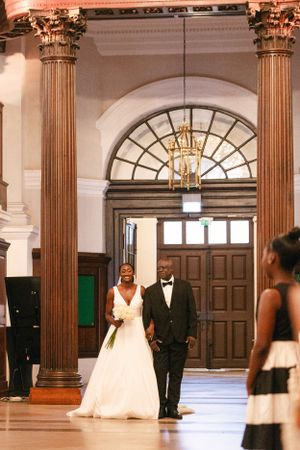 A wedding couple stands beneath ornate classical columns in an elegant venue hallway.