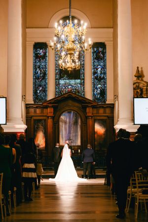 A bride and groom stand at the altar of an elegant church with stained glass windows and a crystal chandelier.