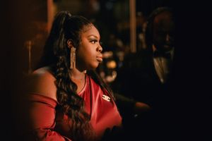 A woman in a red top with long braided hair looks to the side in dramatic lighting.