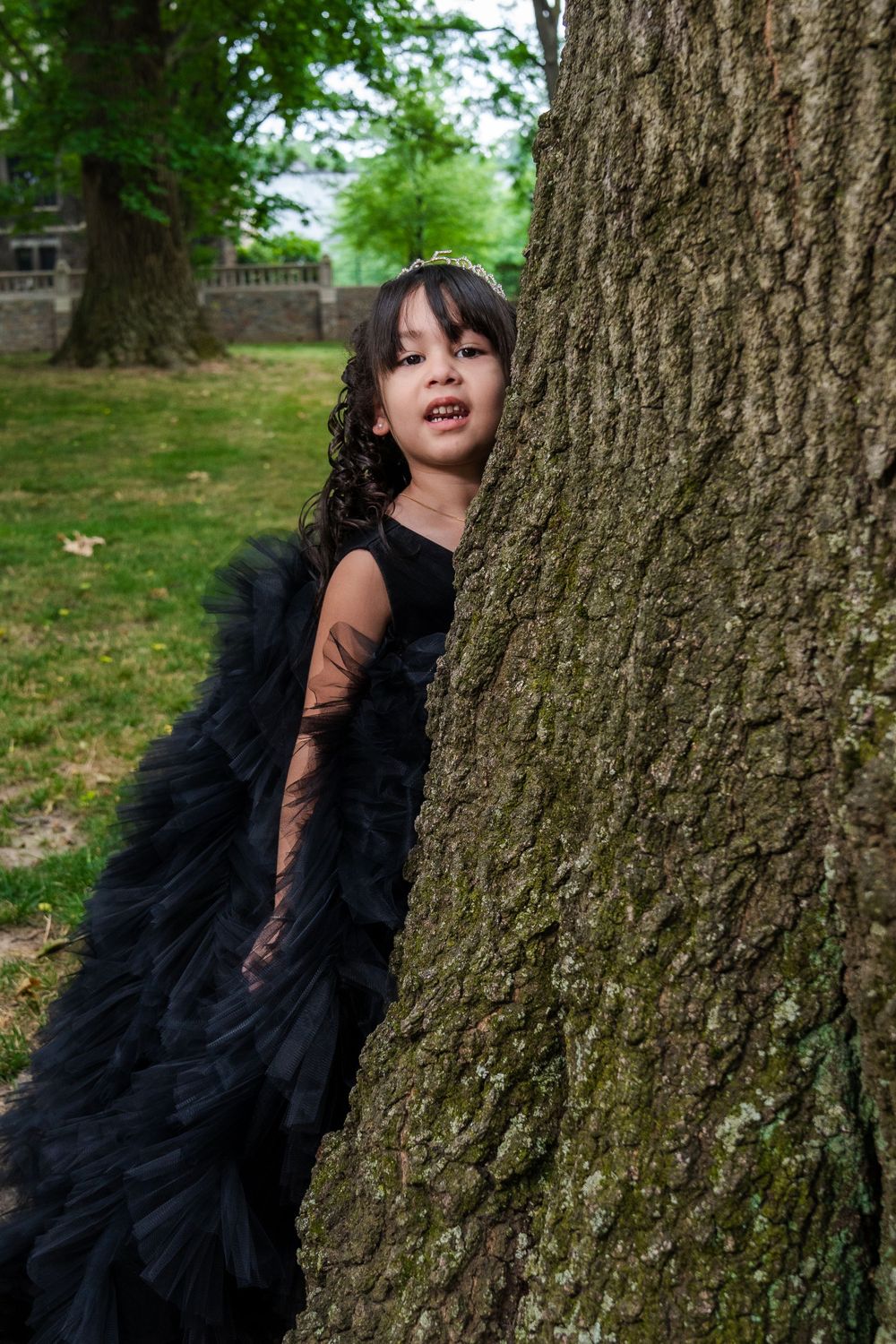 A child in a dramatic black dress peeks out from behind a large tree trunk in a park setting.