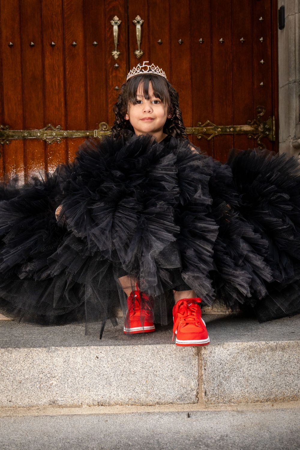 A child wearing a fancy black tulle dress and red sneakers sits on stone steps by a wooden door.