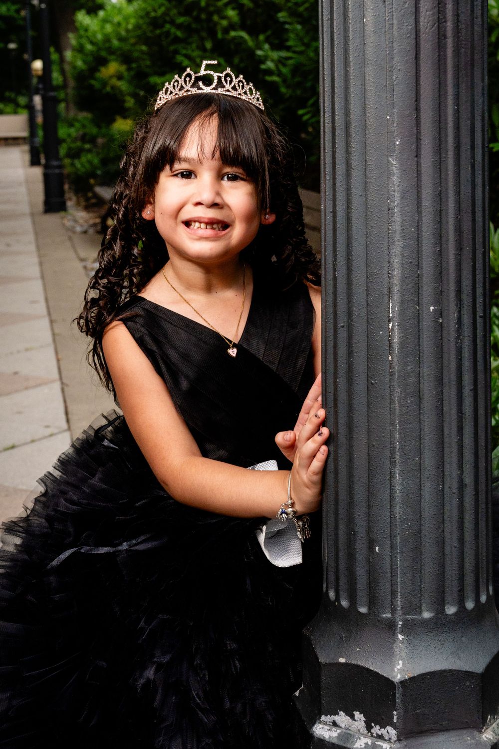 A child in a black tulle dress and tiara poses playfully against a column outdoors.