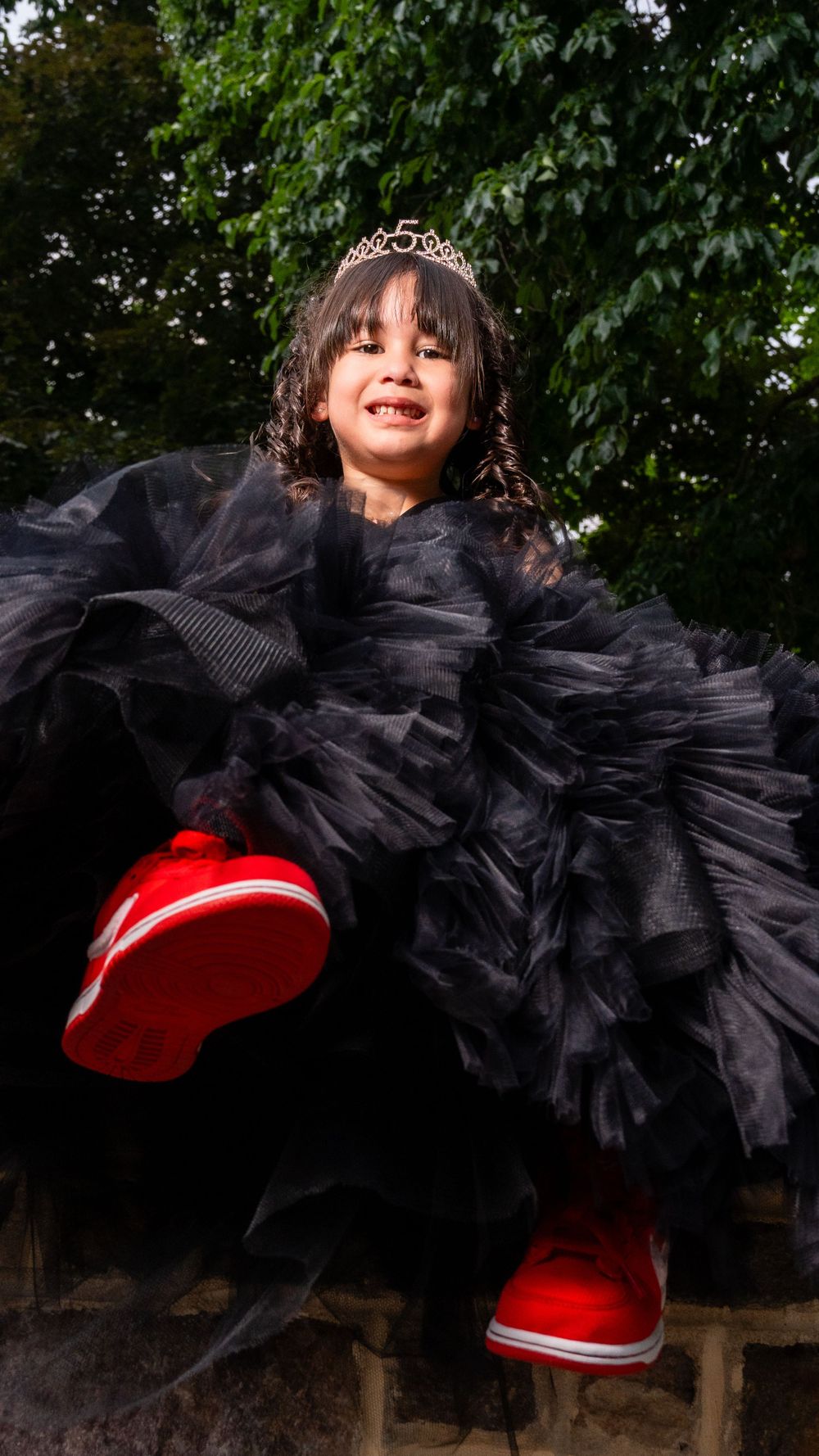 A joyful portrait of a child in a black tulle dress and red shoes jumping outdoors.