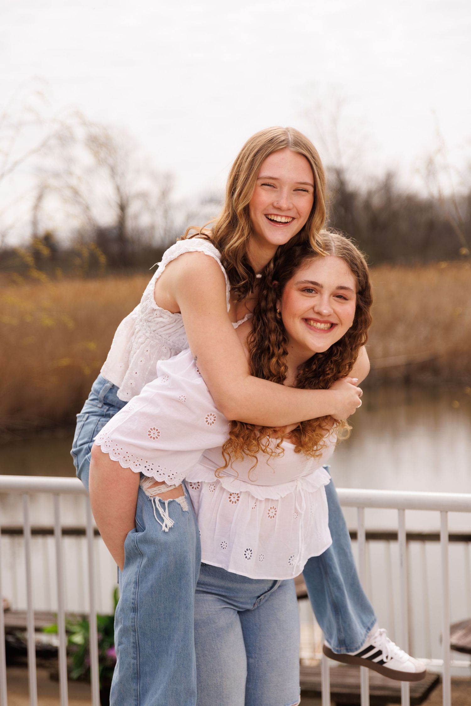 Best friends sharing a playful piggyback ride moment by the lake, wearing matching white tops and jeans.