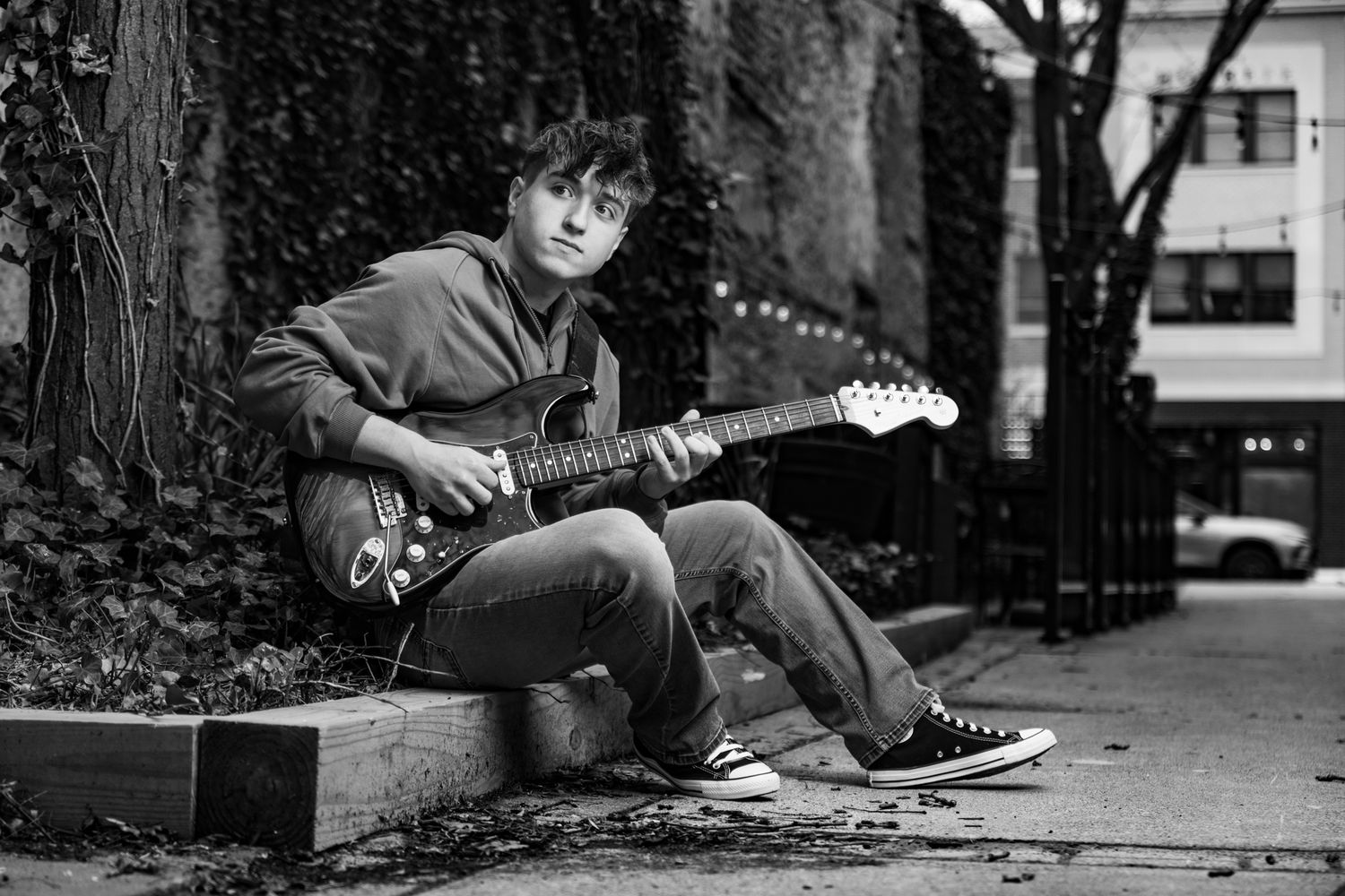 Black and white portrait of musician playing electric guitar while sitting on urban curb near trees.