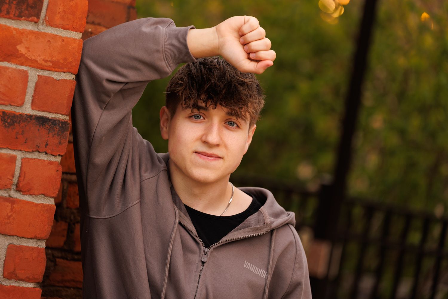 A person in a brown hoodie stands against a brick wall with an autumn forest backdrop.