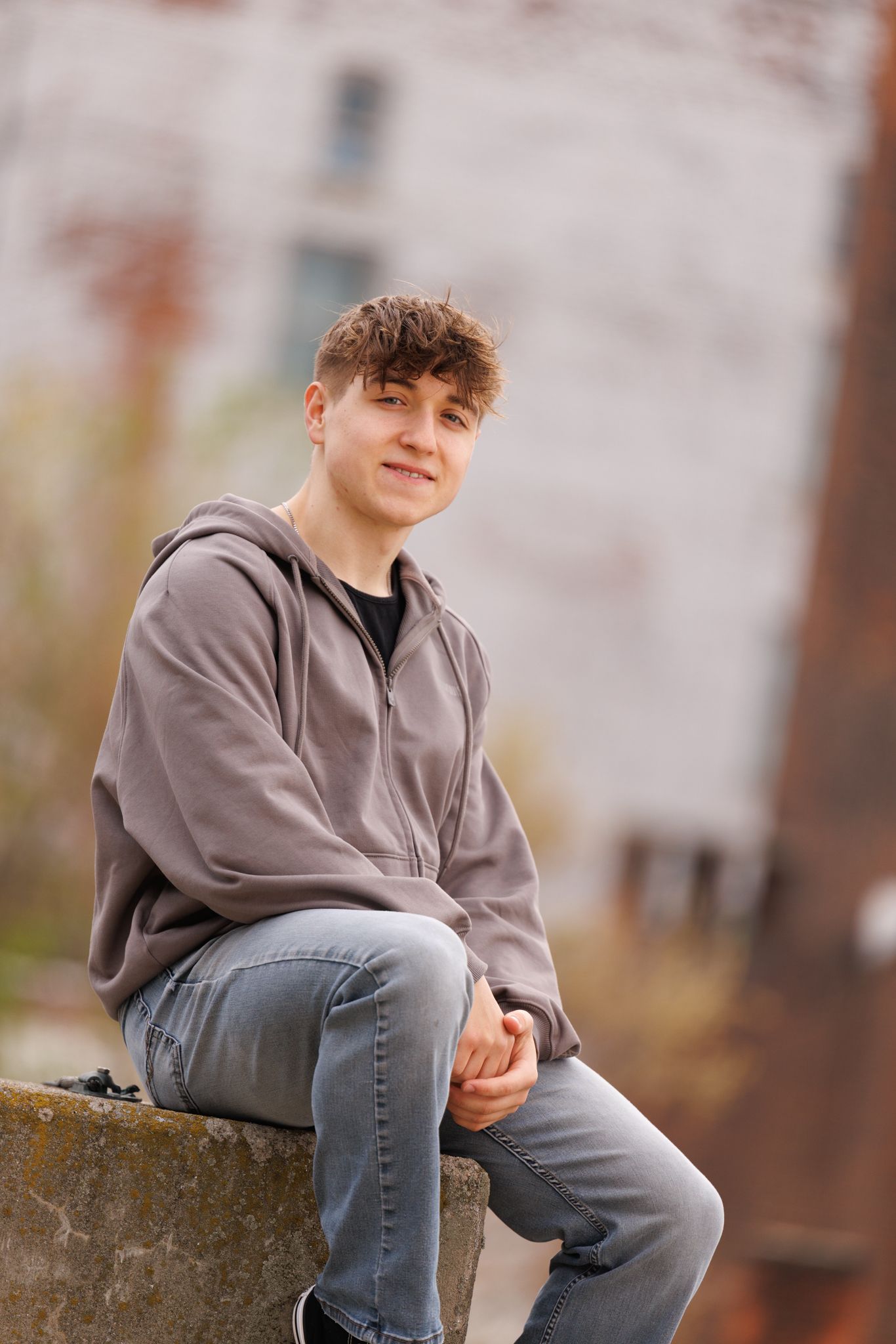 Teen wearing gray hoodie and jeans sits casually on stone ledge during outdoor portrait photo session.