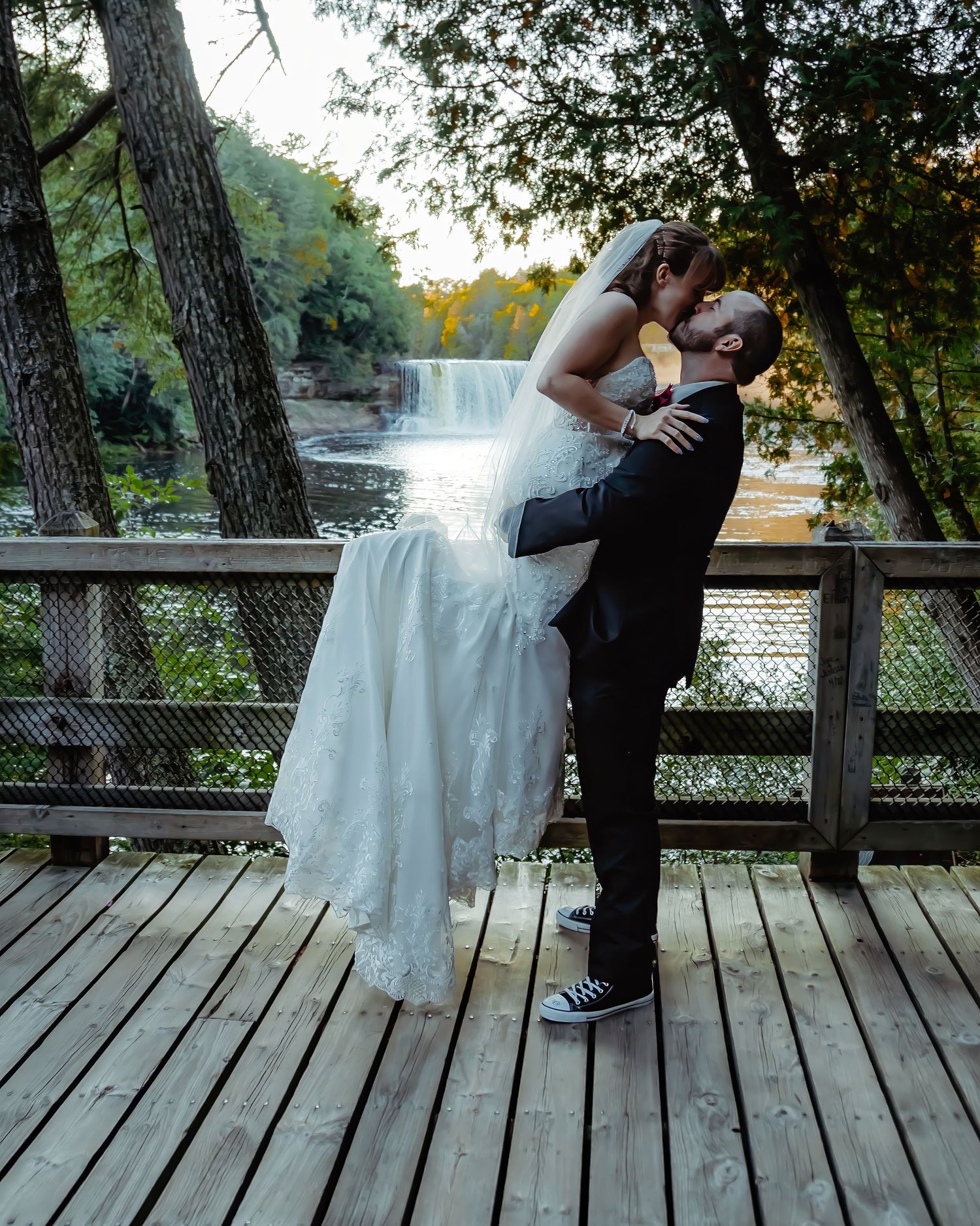 Romantic wedding couple embracing on wooden deck overlooking waterfall at sunset.