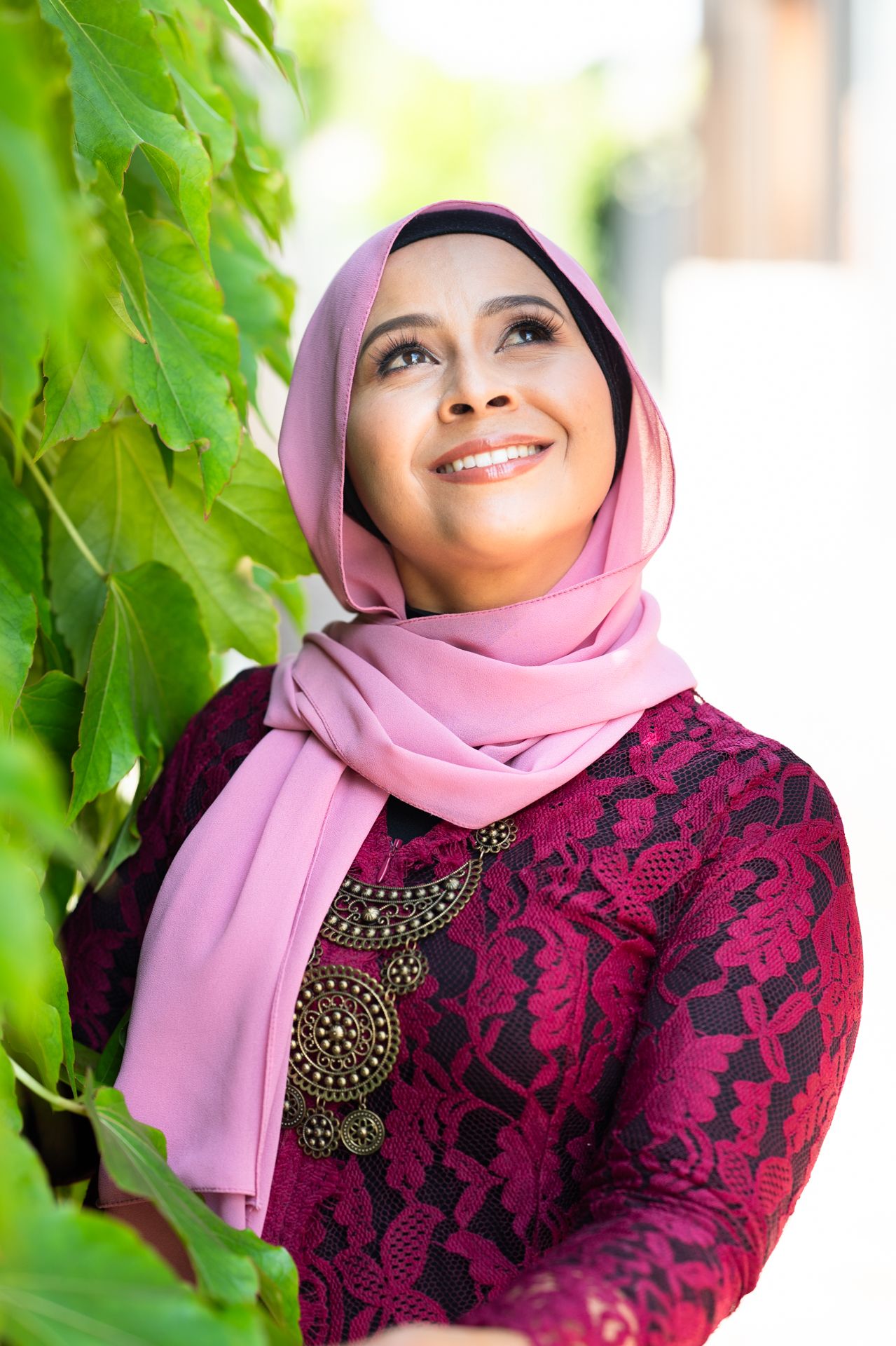 Smiling woman in pink hijab and red lace dress looks upward while standing near lush green leaves outdoors.