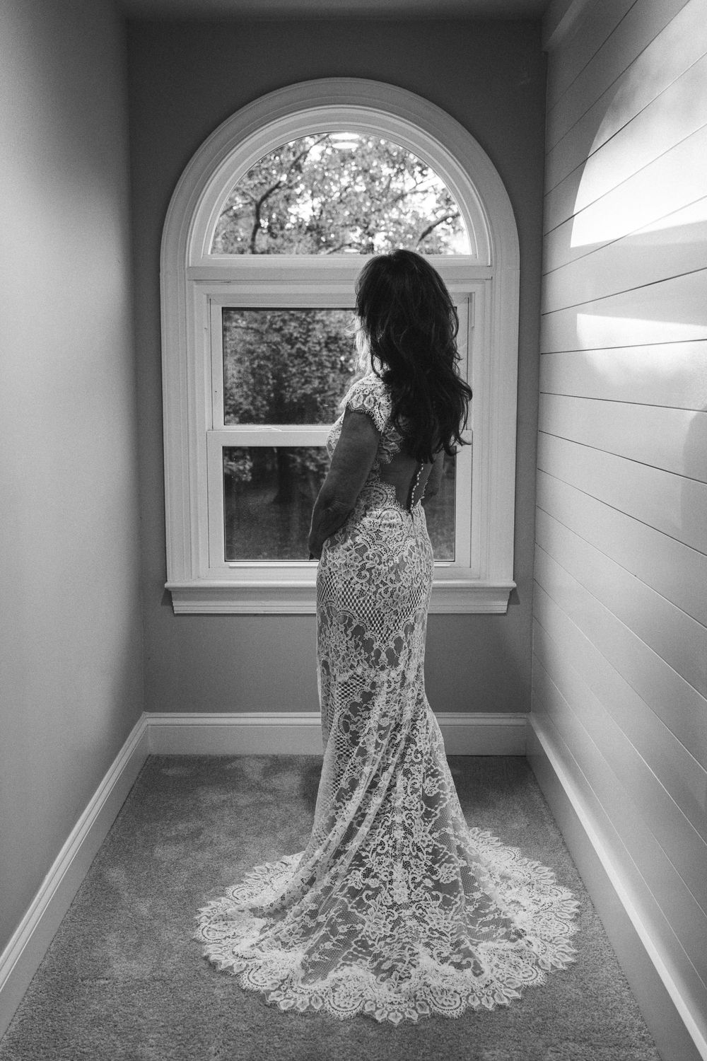 A silhouetted figure in an elegant lace wedding dress stands before an arched window in a hallway.