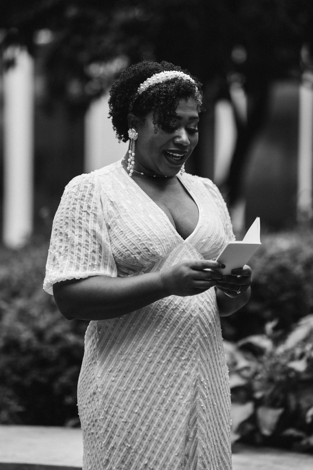 A bride in a white crochet dress reads from a card during an outdoor wedding ceremony.