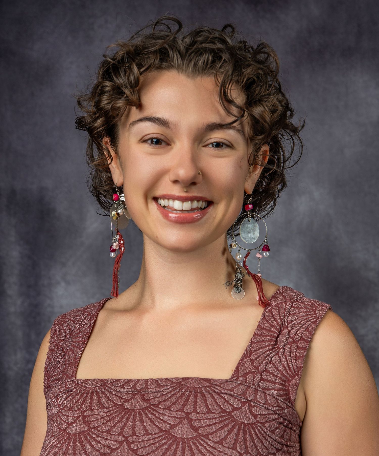 Studio portrait showing curly hair, dangling earrings, and a burgundy lace top against a gray backdrop. Business headshot, woman owned
