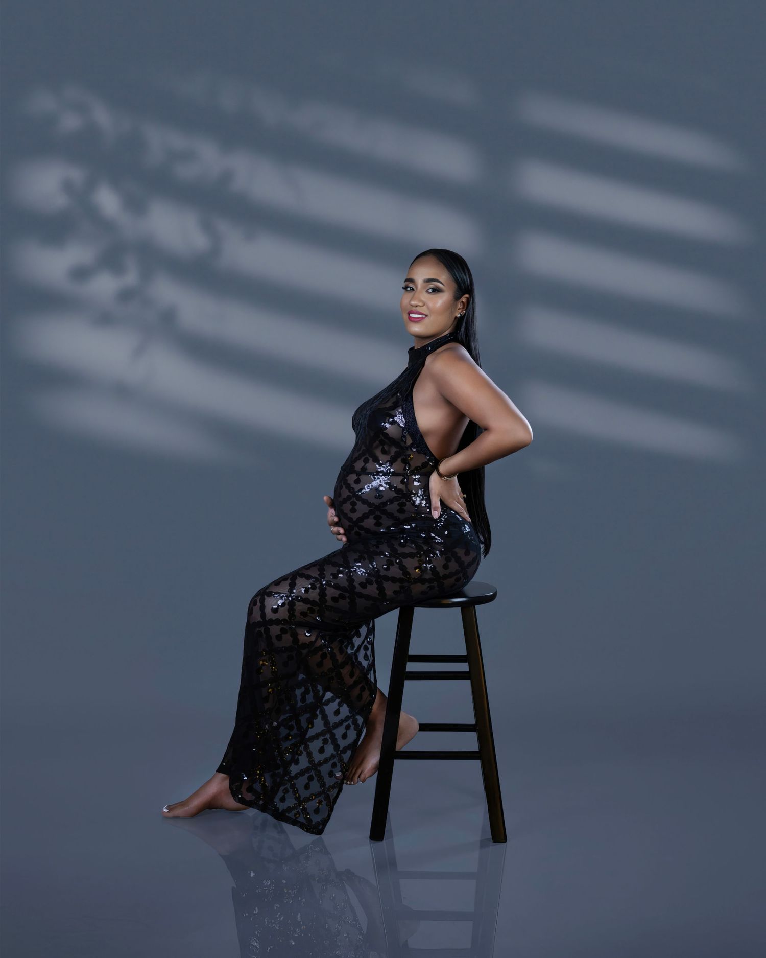 A person in an elegant black lace gown poses gracefully on a wooden stool against a moody gray backdrop.