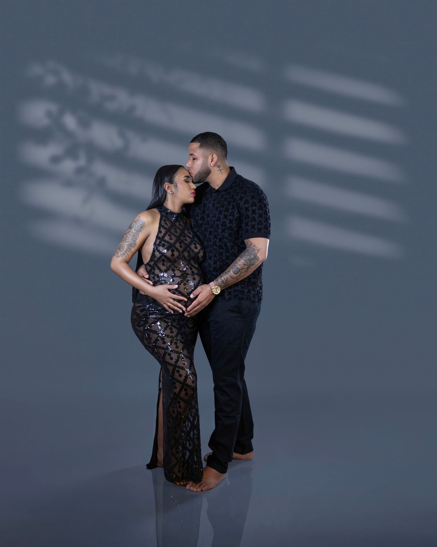 Couple embraces in intimate maternity photo shoot wearing black outfits against gray backdrop with dramatic lighting.