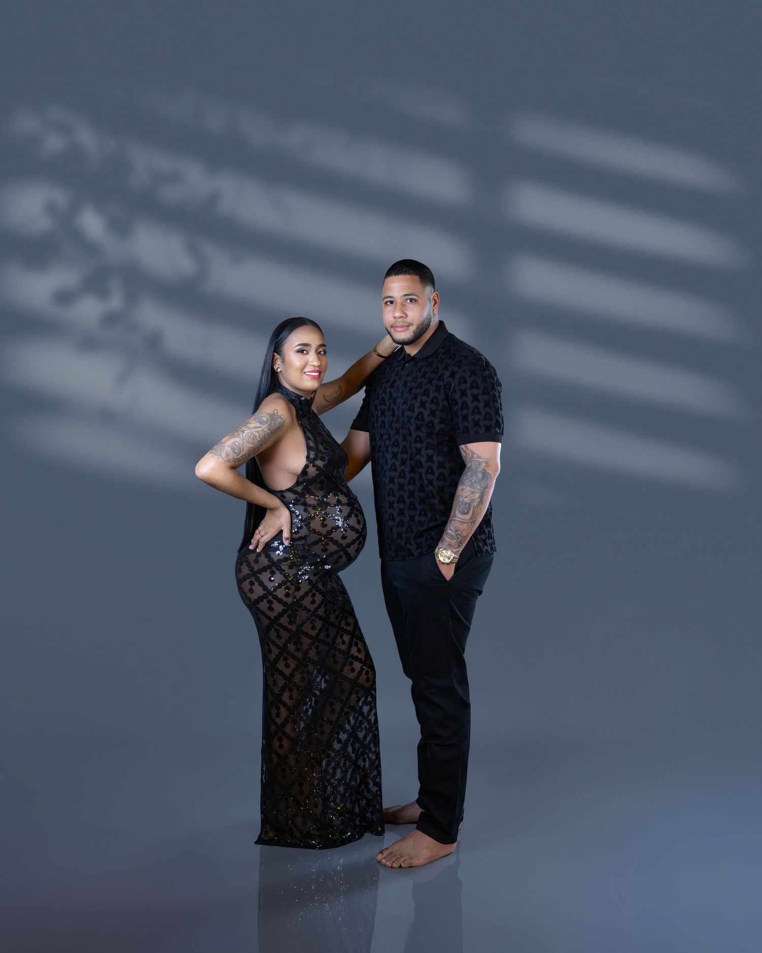 A couple poses together against a dark backdrop with dramatic lighting, wearing elegant evening attire.