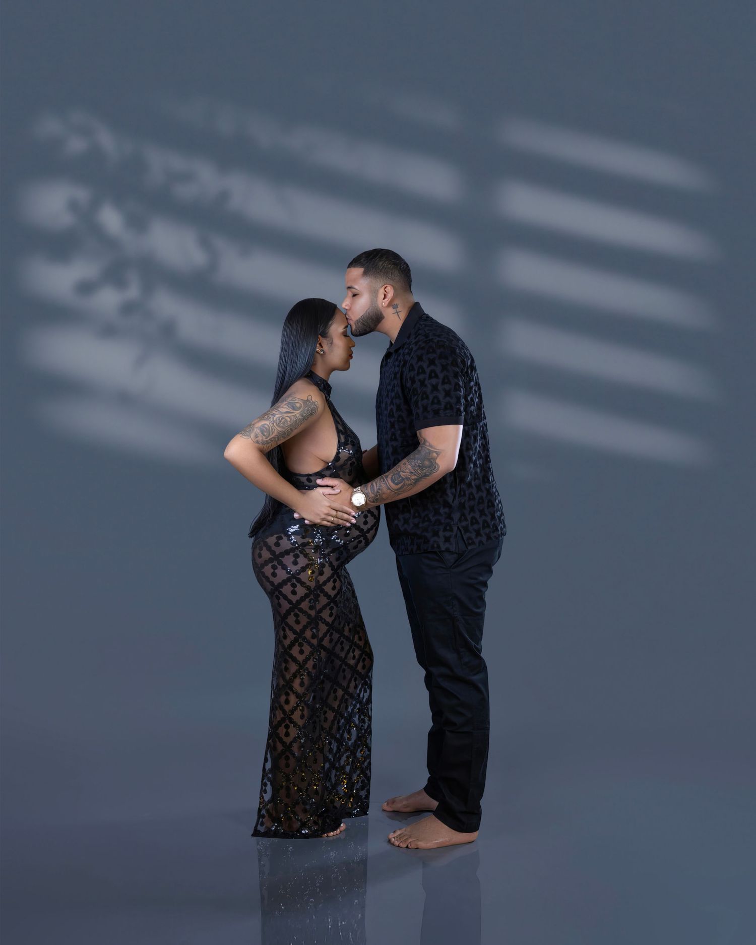 A couple in elegant attire embraces tenderly against a dark backdrop with dramatic window light casting shadows.