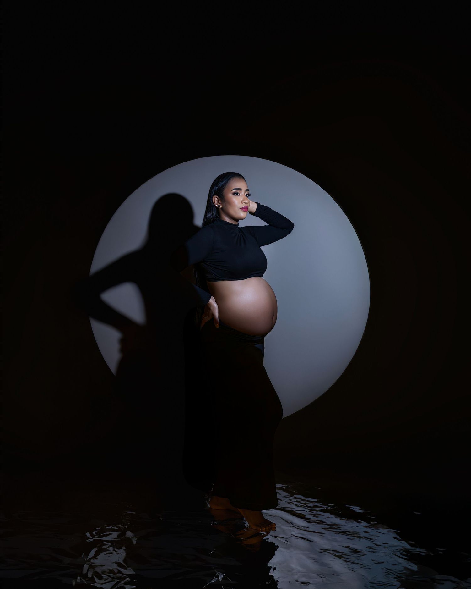 A moody silhouette maternity photo with a circular light backdrop and shadow cast on a wet reflective surface.