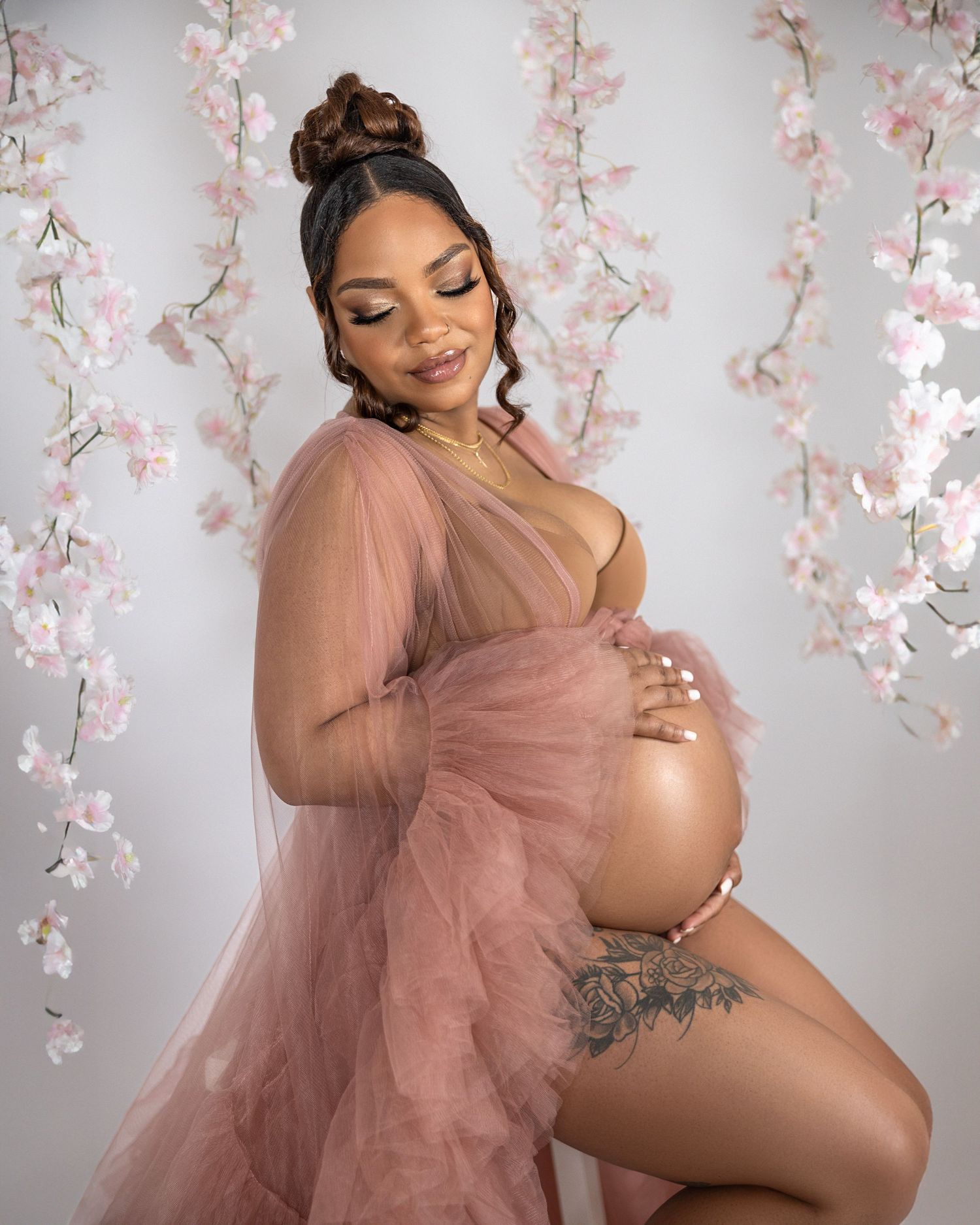 An elegant maternity portrait shows a glowing mom-to-be in a flowing pink tulle gown surrounded by cherry blossoms.
