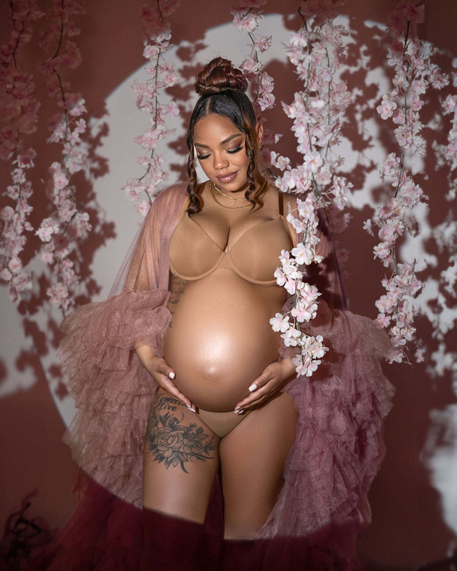 Artistic maternity photo with flowing pink tulle robe and delicate white cherry blossoms against burgundy backdrop.