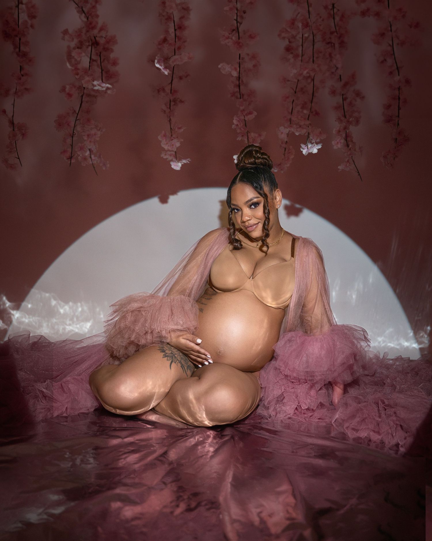 Artistic maternity photo featuring pink tulle fabric and hanging floral elements against a dark burgundy backdrop.