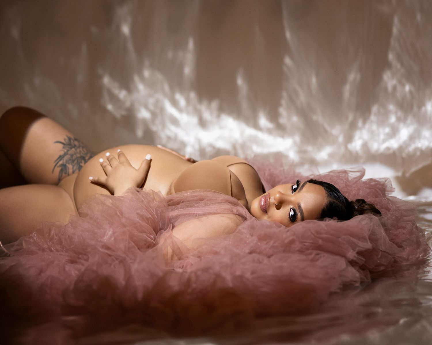 An artistic boudoir photo featuring a feminine figure reclining on pink tulle fabric with dramatic lighting effects.