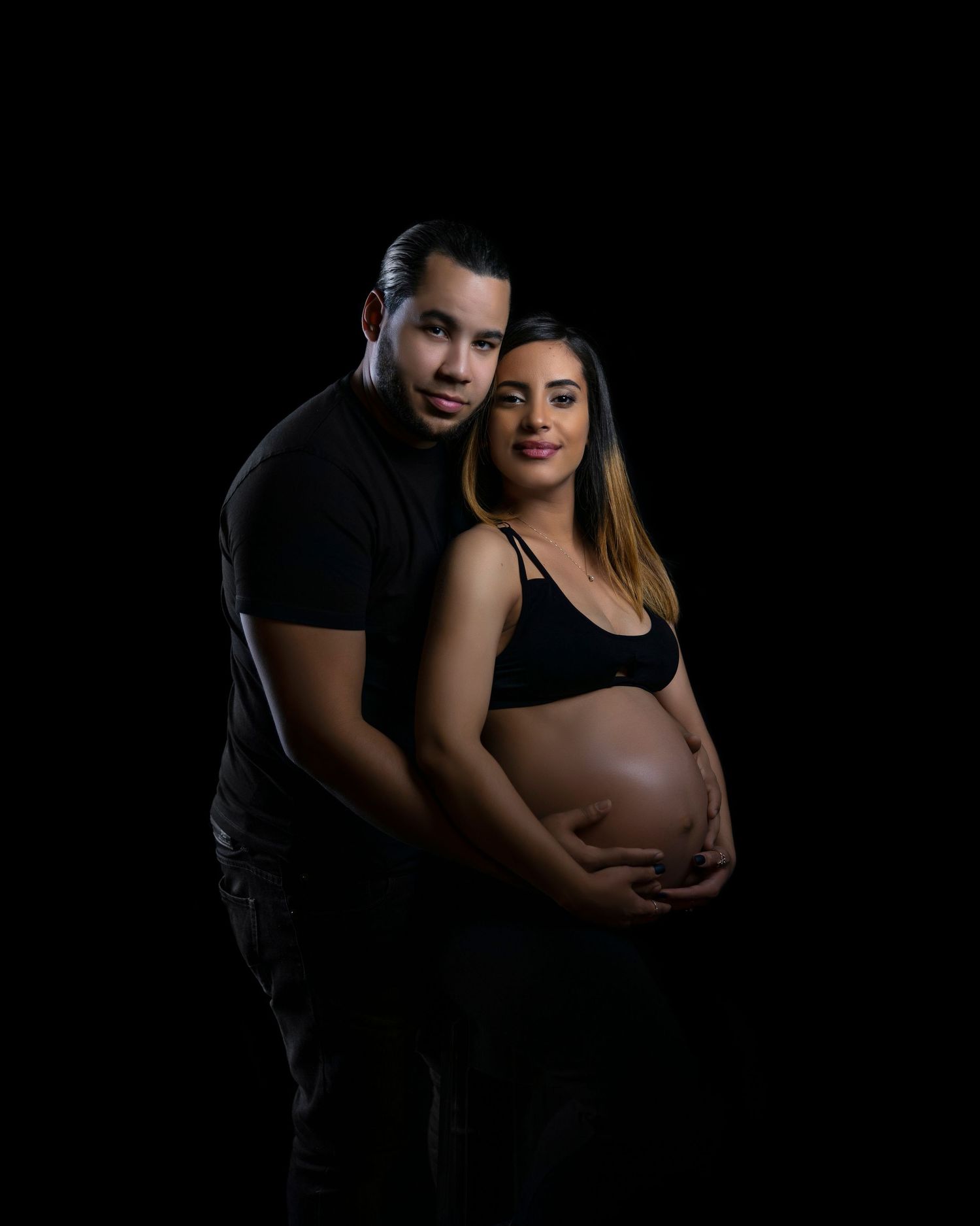 A maternity couple in black outfits pose against a dark background in an intimate studio portrait.