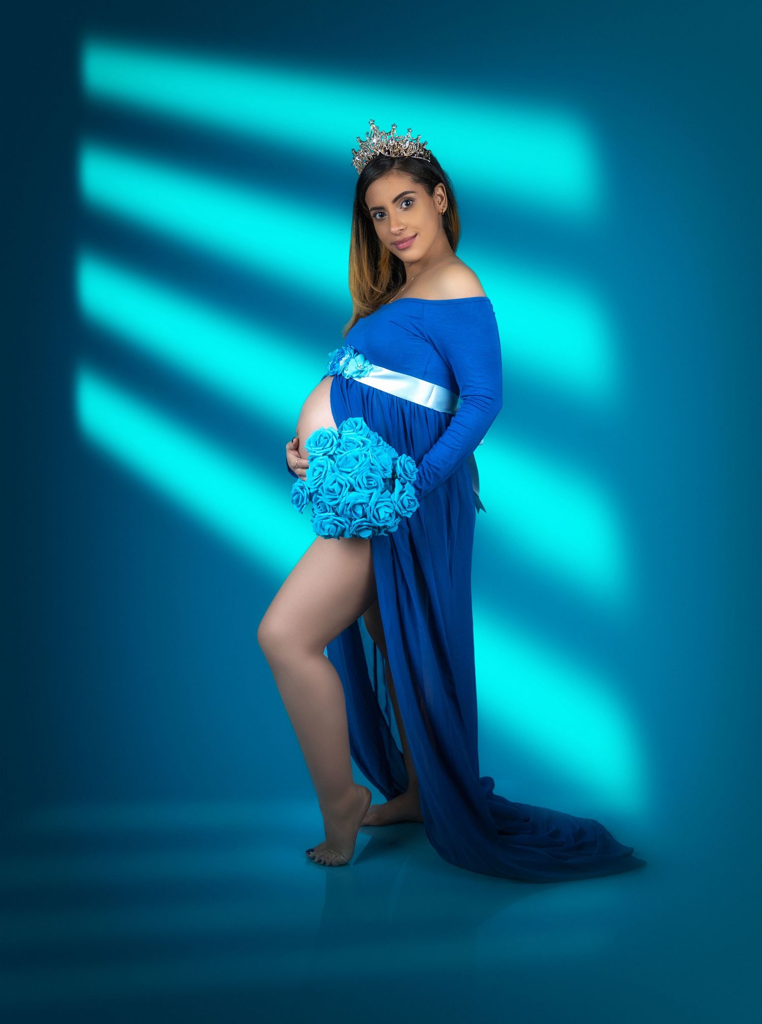 A maternity photoshoot with a royal blue gown and tiara against a turquoise backdrop with dramatic lighting effects.