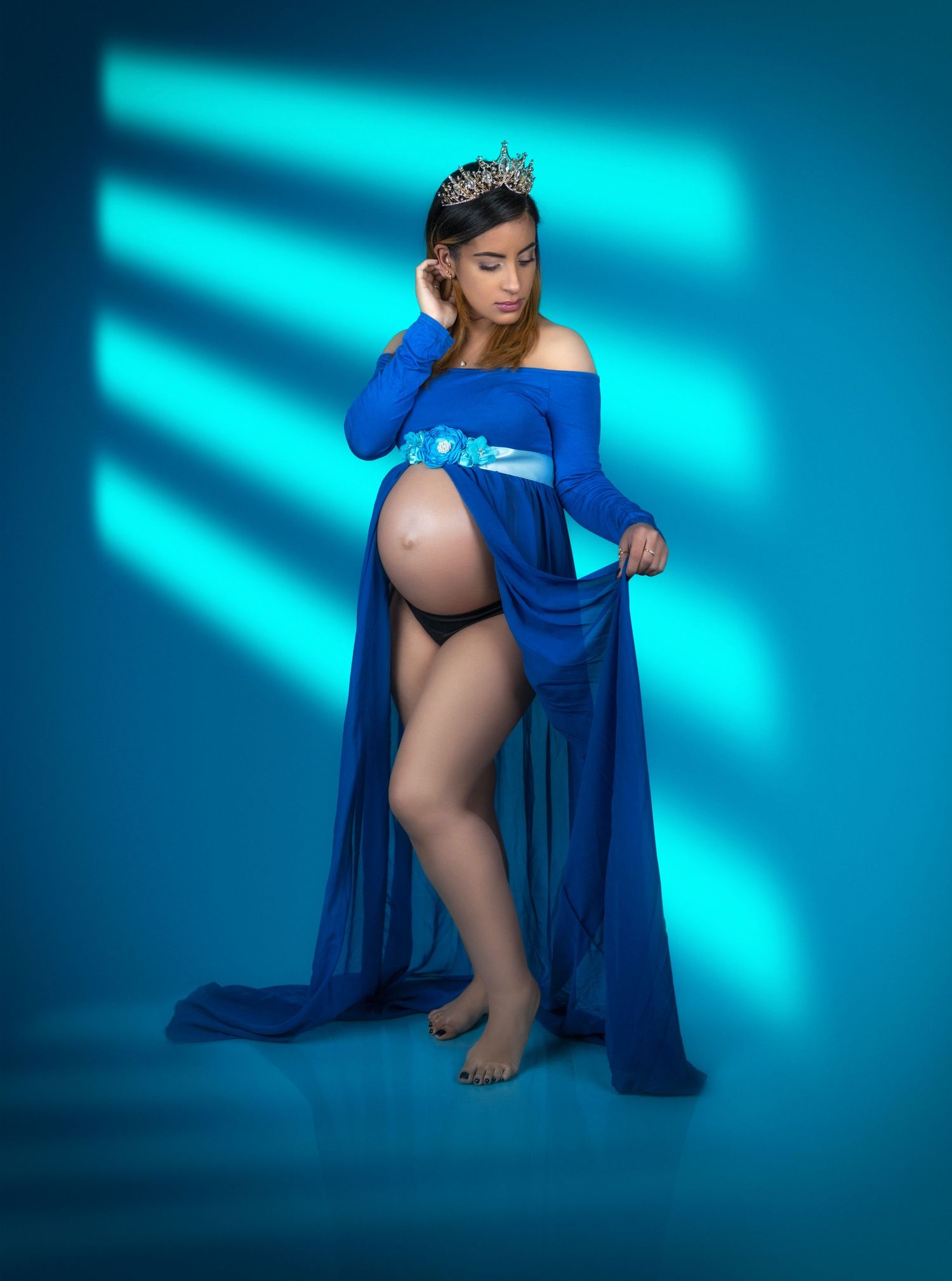 A pregnant woman in a flowing royal blue off-shoulder gown poses against a turquoise-lit backdrop during a maternity photoshoot.