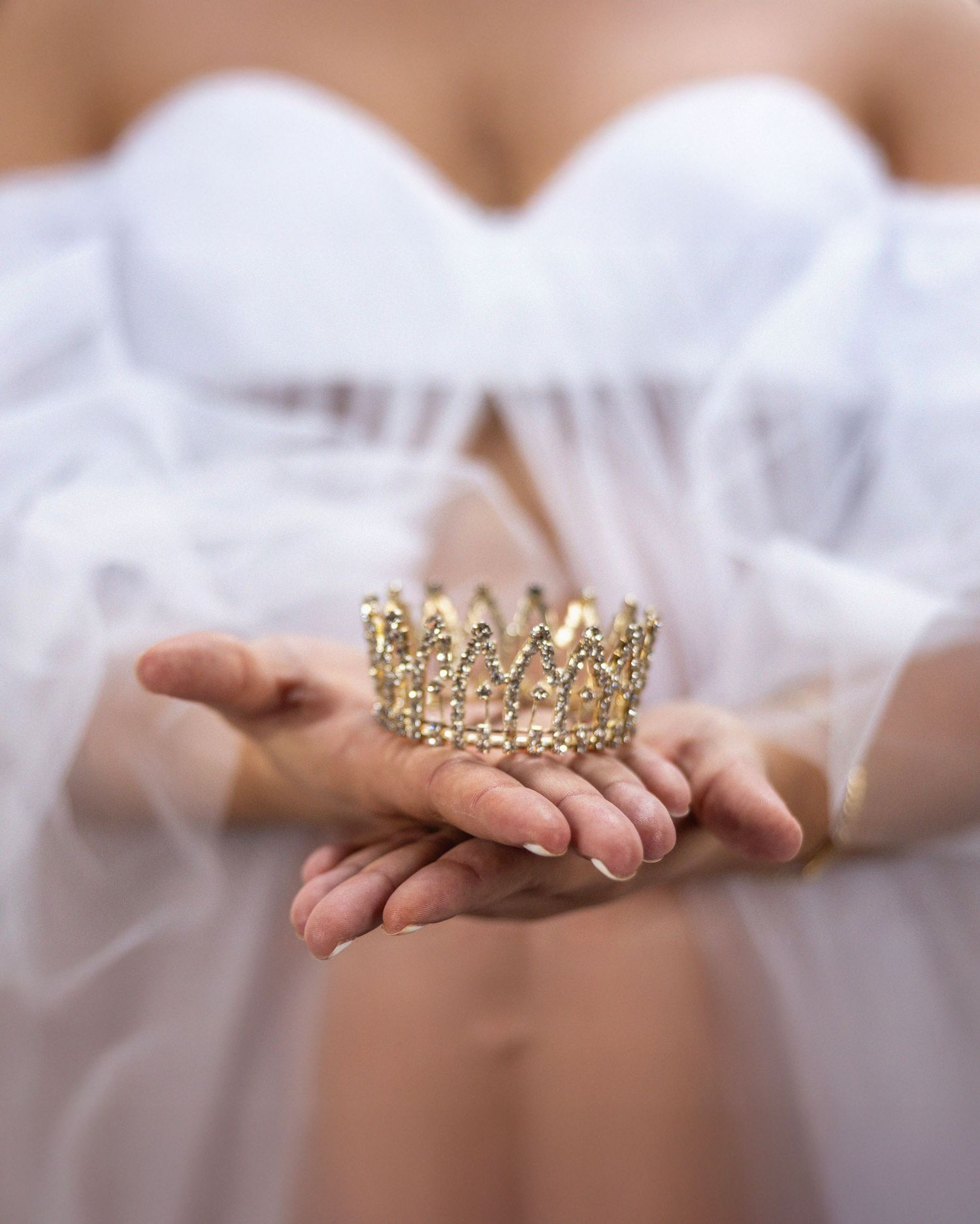 A delicate gold crown held in hands against flowing white tulle fabric, creating an ethereal bridal scene.