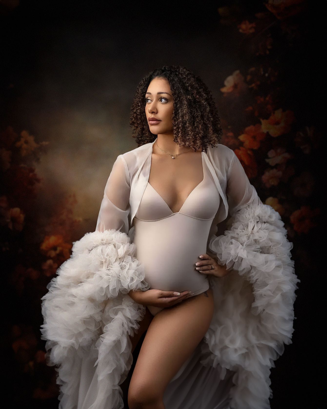 A dreamy maternity portrait in moody lighting features a white bodysuit and flowing sheer robe against warm autumn colors.